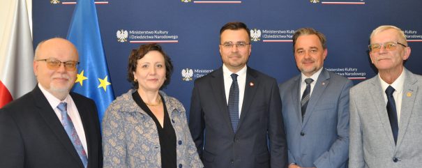 Pięć osób ustawionych w rzędzie na tle granatowego baneru z powtarzającym się napisem „Ministerstwo Kultury i Dziedzictwa Narodowego” i godłem orła, obok widoczne flagi Polski i Unii Europejskiej; trzech mężczyzn w ciemnych garniturach i krawatach, kobieta w jasnej wzorzystej marynarce oraz starszy mężczyzna w jasnoszarym garniturze i okularach.