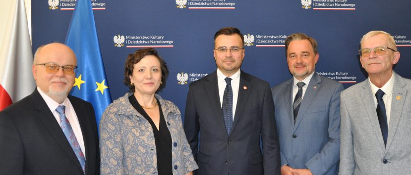 Pięć osób ustawionych w rzędzie na tle granatowego baneru z powtarzającym się napisem „Ministerstwo Kultury i Dziedzictwa Narodowego” i godłem orła, obok widoczne flagi Polski i Unii Europejskiej; trzech mężczyzn w ciemnych garniturach i krawatach, kobieta w jasnej wzorzystej marynarce oraz starszy mężczyzna w jasnoszarym garniturze i okularach.