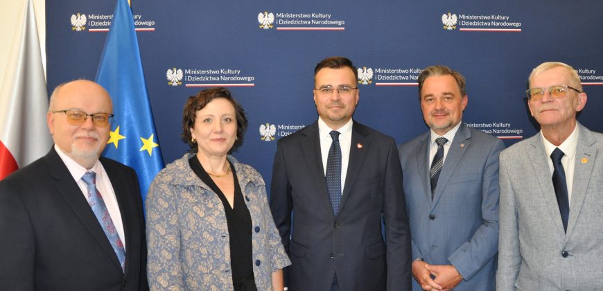 Pięć osób ustawionych w rzędzie na tle granatowego baneru z powtarzającym się napisem „Ministerstwo Kultury i Dziedzictwa Narodowego” i godłem orła, obok widoczne flagi Polski i Unii Europejskiej; trzech mężczyzn w ciemnych garniturach i krawatach, kobieta w jasnej wzorzystej marynarce oraz starszy mężczyzna w jasnoszarym garniturze i okularach.