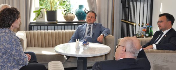 Kilka osób siedzi na beżowych pikowanych kanapach w jasnym salonie wokół okrągłego marmurowego stolika; w tle okno z doniczkowymi roślinami i dekoracyjnymi wazonami, obok lampa stojąca i konsola z ramkami zdjęć oraz butelkami i szklankami.