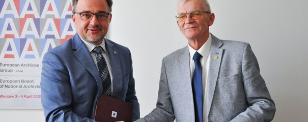 Dwóch mężczyzn w garniturach stoi na tle białej ściany i plakatu z powtarzającym się motywem litery A; po lewej mężczyzna w niebieskim garniturze i okularach trzyma bordową teczkę, po prawej starszy mężczyzna w jasnoszarym garniturze i okularach trzyma granatową teczkę, obaj ściskają dłonie.