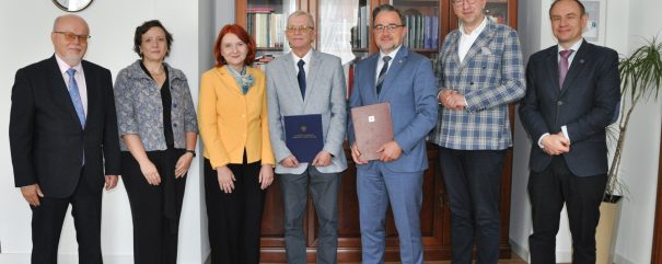 Siedem dorosłych osób ustawionych w rzędzie przed dużą drewnianą szafą z przeszklonymi półkami pełnymi książek; większość w formalnych strojach (m.in. kobieta w żółtej marynarce z apaszką), dwie osoby trzymają teczki z dokumentami, po lewej na ścianie okrągły zegar, po prawej wysoka doniczka z rośliną i drewniana podłoga.