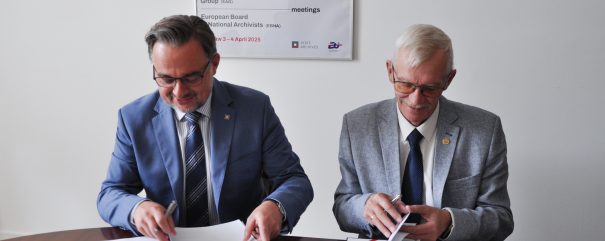 Dwaj mężczyźni w okularach i garniturach siedzą przy drewnianym stole, trzymając długopisy i przewracając dokumenty do podpisania, a za nimi na ścianie wisi plakat z powtarzającymi się kolorowymi literami A i napisem European Archives Group