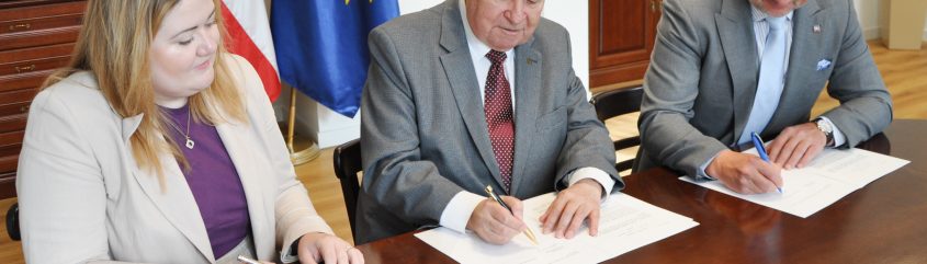 Trzy osoby siedzą przy drewnianym stole podpisując dokumenty: po lewej kobieta z jasnymi włosami w jasnej marynarce i fioletowej bluzce, pośrodku mężczyzna z przerzedzonymi włosami w popielatym garniturze i krawacie w kropki podpisujący piórem, po prawej mężczyzna w okularach i garniturze z jasnym krawatem podpisujący długopisem; w tle stoją flagi Polski i Unii Europejskiej, regał z książkami i roślina, na stole widoczne pudełko z piórem.