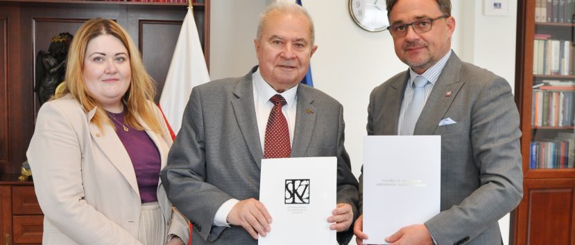 Trzech stojących dorosłych osób ujętych w półpostaci — kobieta w beżowej marynarce i fioletowym topie po lewej, starszy mężczyzna w szarej marynarce i czerwonym krawacie pośrodku oraz mężczyzna w szarej marynarce, okularach i jasnoniebieskim krawacie po prawej — trzymają białe teczki z dokumentami; w tle drewniane regały z książkami i rzeźbą, flagi i okrągły zegar na ścianie.