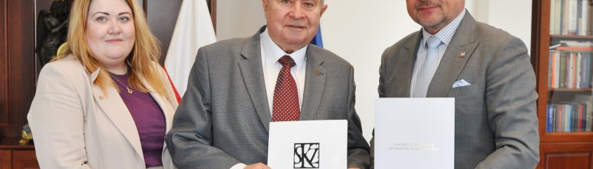 Trzech stojących dorosłych osób ujętych w półpostaci — kobieta w beżowej marynarce i fioletowym topie po lewej, starszy mężczyzna w szarej marynarce i czerwonym krawacie pośrodku oraz mężczyzna w szarej marynarce, okularach i jasnoniebieskim krawacie po prawej — trzymają białe teczki z dokumentami; w tle drewniane regały z książkami i rzeźbą, flagi i okrągły zegar na ścianie.