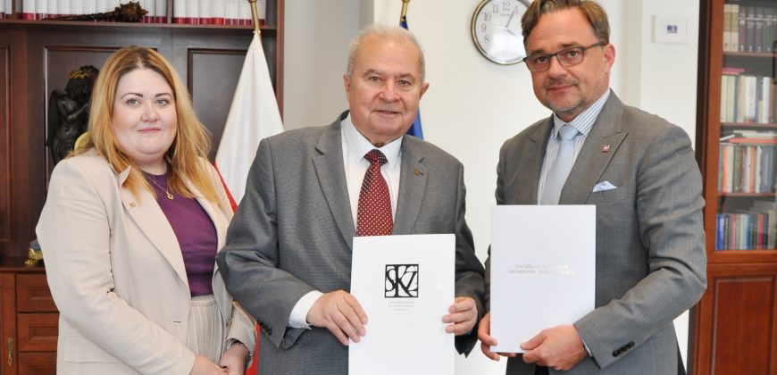 Trzech stojących dorosłych osób ujętych w półpostaci — kobieta w beżowej marynarce i fioletowym topie po lewej, starszy mężczyzna w szarej marynarce i czerwonym krawacie pośrodku oraz mężczyzna w szarej marynarce, okularach i jasnoniebieskim krawacie po prawej — trzymają białe teczki z dokumentami; w tle drewniane regały z książkami i rzeźbą, flagi i okrągły zegar na ścianie.