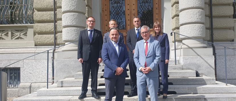 Sześć osób w formalnych strojach (marynarki, krawaty) stoi na szerokich kamiennych schodach przed ozdobnymi drewnianymi drzwiami z kutymi metalowymi kratami, po bokach widoczne masywne zaokrąglone rustykalne kolumny i zdobione okna fasady.