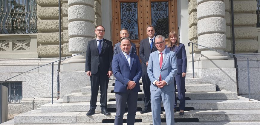 Sześć osób w formalnych strojach (marynarki, krawaty) stoi na szerokich kamiennych schodach przed ozdobnymi drewnianymi drzwiami z kutymi metalowymi kratami, po bokach widoczne masywne zaokrąglone rustykalne kolumny i zdobione okna fasady.