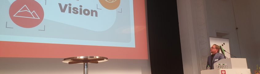 Scena sali konferencyjnej z dużym, częściowo widocznym slajdem po lewej z napisem 