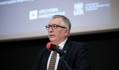 Mężczyzna w okularach i siwych włosach przy mikrofonie z czerwonym filtrem, ubrany w ciemny garnitur, białą koszulę i wzorzysty krawat, z przypinką na klapie i długopisem w kieszeni, na tle rozmytego ekranu projekcji z białym tekstem i logotypami.