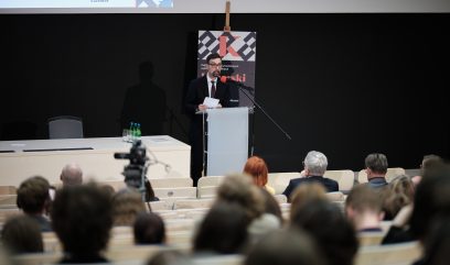 Prelegent w ciemnym garniturze i okularach stoi przy przezroczystym pulpicie z mikrofonem i trzyma kartki, za nim na stojaku plakat z dużą literą K, w przodzie sala wykładowa z rozmytymi uczestnikami na widowni, stołem z butelkami wody i kamerą nagrywającą.