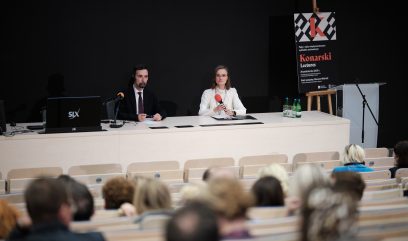 Na scenie sala wykładowa: dwóch prelegentów — mężczyzna w ciemnym garniturze z mikrofonem na stojaku i słuchawką oraz kobieta w jasnej bluzce w kropki i okularach trzymająca czerwony mikrofon, siedzą za długim jasnym stołem z laptopem, butelkami wody i dokumentami, na ciemnym tle po prawej stoi plakat z napisem 