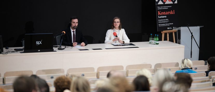 Na scenie sala wykładowa: dwóch prelegentów — mężczyzna w ciemnym garniturze z mikrofonem na stojaku i słuchawką oraz kobieta w jasnej bluzce w kropki i okularach trzymająca czerwony mikrofon, siedzą za długim jasnym stołem z laptopem, butelkami wody i dokumentami, na ciemnym tle po prawej stoi plakat z napisem 