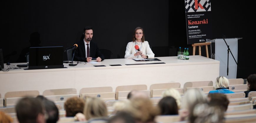 Na scenie sala wykładowa: dwóch prelegentów — mężczyzna w ciemnym garniturze z mikrofonem na stojaku i słuchawką oraz kobieta w jasnej bluzce w kropki i okularach trzymająca czerwony mikrofon, siedzą za długim jasnym stołem z laptopem, butelkami wody i dokumentami, na ciemnym tle po prawej stoi plakat z napisem 