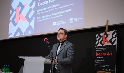 Mężczyzna w okularach i szarym garniturze stoi przy przezroczystej mównicy z mikrofonem, na tle dużego ekranu z projekcją plakatu 