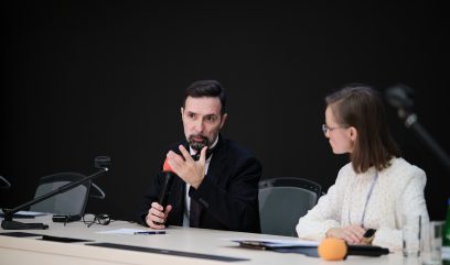 Mężczyzna w ciemnym garniturze z krótkimi włosami i brodą trzyma mikrofon i gestykuluje, obok niego siedzi kobieta w białej bluzce i okularach; oboje są przy stole z dokumentami, okularami, mikrofonami (w tym pomarańczową osłoną) oraz butelką wody na tle ciemnej ściany.
