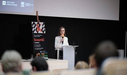 Osoba w jasnej bluzce z mikrofonem stoi za białą mównicą na scenie przed czarnym tłem, obok na sztaludze plakat z dużym napisem Konarski Lectures, nad sceną widoczny duży biały ekran z fragmentem slajdu i logo Archiwa Państwowe, a w pierwszym planie na stole leżą butelki wody i rozmazane sylwetki publiczności.