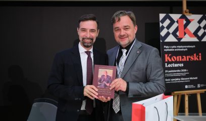 Dwaj mężczyźni w garniturach, obaj uśmiechnięci, stoją na ciemnym tle i razem trzymają bordową książkę z reprodukcją portretu, po prawej widoczny jest stojak z plakatem „Konarski Lectures”, a z przodu biało‑czerwona torba upominkowa.