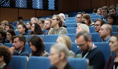 Duży kadr sali wykładowej z wieloma rzędami niebieskich foteli, w których siedzi grupa dorosłych uczestników — większość w okularach, niektórzy z słuchawkami i identyfikatorami — wszyscy zwróceni w stronę przodu, a w tle drewniane panele i okna z perforowanym wzorem.