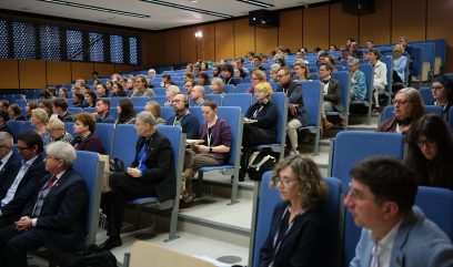 Widok z boku na dużą salę wykładową z wieloma osobami siedzącymi w niebieskich, tarasowo ustawionych fotelach, drewnianymi panelami na ścianach, sufitowym oświetleniem, widocznym rzutnikiem i kilkoma uczestnikami ze słuchawkami lub notatnikami.