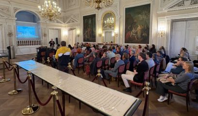 Wnętrze bogato zdobionej sali z kryształowymi żyrandolami i dużymi obrazami, w której publiczność siedzi w rzędach krzeseł oglądając prezentację na ekranie; na pierwszym planie długi oszklony eksponat z ręcznym pismem zabezpieczony sznurkami i słupkami, u frontu stoi osoba przemawiająca, a inna osoba w żółto-czarnej bluzie stoi tyłem.