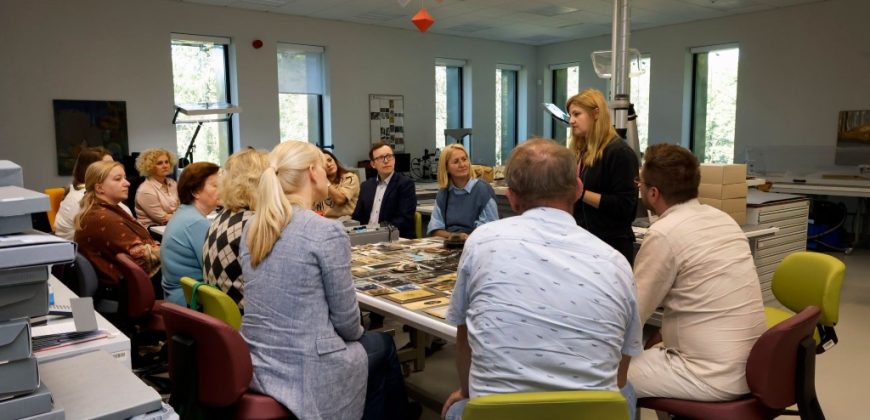 Grupa osób siedzi wokół dużego stołu z rozłożonymi fotografiami i dokumentami, a stojąca kobieta zwraca się do nich; w jasnym pomieszczeniu widać duże okna, kolorowe krzesła, pudełka i wiszącą dekorację sufitową.