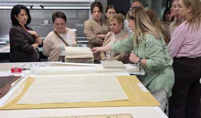 Kilka kobiet stoi przy dużym stole w pomieszczeniu; kobieta w zielonej koszuli podnosi kartę ze stosu starych dokumentów, na stole rozłożone arkusze papieru, żółta teczka i fragment mapy, w tle widać regały i urządzenia.