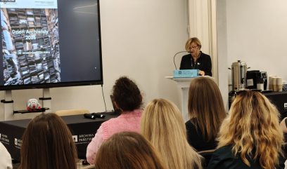 Wnętrze sali konferencyjnej: po lewej duży ekran z slajdem „Dzień Archiwisty 2026 29–30 września 2026 roku” i zdjęciem regałów z aktami, po prawej kobieta w okularach stojąca przy mównicy z mikrofonem, na pierwszym planie widoczne od tyłu głowy kilku uczestniczek, na stole czarny obrus z logo „Archiwa Państwowe”, a w tle ekspres do kawy i kubki.