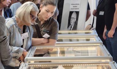 Grupa osób w pomieszczeniu, dwie kobiety na pierwszym planie pochylone nad podświetlanymi przeszklonymi witrynami wystawowymi z dokumentami, w tle plakat z czarno-białym portretem i napisem 