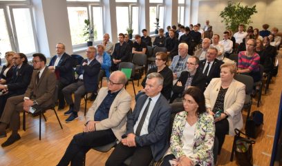 Grupa kilkudziesięciu osób — dorośli i młodzież — siedzi w rzędach krzeseł w jasnej sali konferencyjnej z dużymi oknami po lewej, drewnianą podłogą i kilkoma roślinami doniczkowymi; część osób ubrana formalnie, część casual.