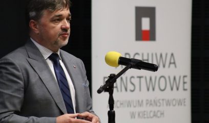Mężczyzna w szarym garniturze, białej koszuli i granatowym krawacie stoi przy mikrofonie na statywie z żółtym filtrem, trzymając ręce przed sobą; z prawej w tle widoczny pionowy biały baner z prostym logo i częściowym napisem 