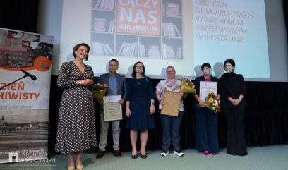 Sześć osób stoi na scenie przed dużym ekranem z grafiką i napisami „ŁĄCZY NAS ARCHIWUM” oraz „OBCHODY DNIA ARCHIWISTY W ARCHIWUM PAŃSTWOWYM W KOSZALINIE”; od lewej kobieta w sukience w groszki, mężczyzna w marynarce trzymający bukiet i dyplom, kobieta w granatowej sukience, kobieta w jasnej bluzce trzymająca oprawioną ramkę, kobieta w granatowym garniturze z bukietem i dyplomem oraz kobieta w czarnym stroju, po lewej stoi baner z napisem „DZIEŃ ARCHIWISTY” i zdjęciem budynku.
