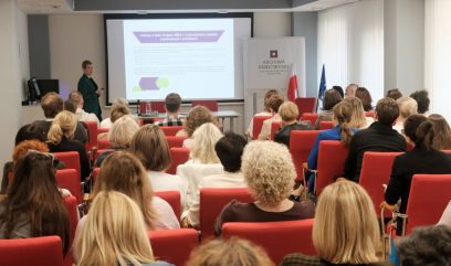 Widok z tyłu na salę konferencyjną z rzędami czerwonych foteli wypełnionych uczestnikami; osoba prowadząca stoi po lewej przy dużym ekranie z wyświetloną prezentacją, obok umieszczony roll‑up z napisem 