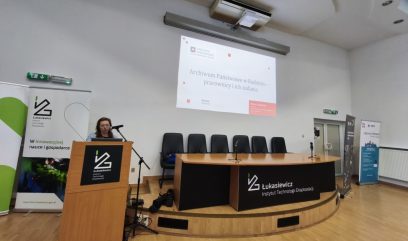 Sala konferencyjna z drewnianą podłogą, na pierwszym planie podium z logo Łukasiewicz, przy którym stoi kobieta przemawiająca do mikrofonu, obok półokrągłe biurko z kilkoma krzesłami i mikrofonami, nad nimi projektor wyświetlający slajd z napisem Archiwum Państwowe w Radomiu – pracownicy i ich zadania oraz stojące roll-upy z logo Łukasiewicz.