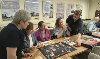 Grupa osób siedzi i stoi przy długim drewnianym stole w jasnym pomieszczeniu z gablotami i planszami informacyjnymi na ścianie, wspólnie grając w rozłożoną na stole planszówkę z planszą, kartami, kostkami i żetonami, podczas gdy jedna osoba kładzie kartę, a pozostali obserwują rozgrywkę.