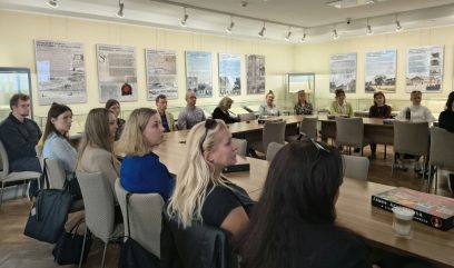 Grupa osób dorosłych siedzi przy rozmieszczonych w kształt litery U drewnianych stołach w sali wystawowej z informacyjnymi planszami i przeszklonymi gablotami na ścianach, oświetlona sufitowymi reflektorami; na stolikach widać torby, kubek z kawą i pudełko gry.