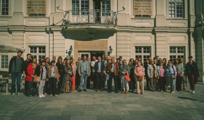 Duża grupa dorosłych osób ustawionych w poziomym szeregu przed jasną klasycystyczną fasadą budynku z balkonem i tablicą 