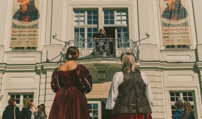 Przed jasną neoklasycystyczną fasadą z ozdobnym balkonem stoi grupa widzów, w tym dwie osoby widziane od tyłu w bordowych, historyzujących sukniach, patrzące na osobę stojącą w otwartym oknie balkonu, a po bokach elewacji wiszą dwa duże plakaty z profilami twarzy.
