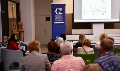 Widok z tyłu sali prezentacyjnej: grupa uczestników siedzi na kolorowych krzesłach zwrócona ku dużemu ekranowi projekcyjnemu, po lewej siedzi prelegentka z mikrofonem, obok stoi pionowy baner z napisem KSIĄŻNICA ZAMOJSKA, a przy ścianie widoczne są dwa portrety na sztalugach.