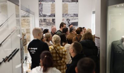 Grupa osób stojąca w wąskim wnętrzu muzeum przed ścianą z historycznymi planszami i opisami, po lewej w gablocie widoczne bagnety, po prawej fragment munduru wojskowego.