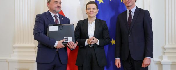 Trzy osoby w formalnych garniturach stoją w ozdobnym białym wnętrzu na tle flagi Polski i flagi Unii Europejskiej; mężczyzna po lewej trzyma czarny futerał z białymi naklejkami, kobieta stoi pośrodku, a mężczyzna po prawej ma okulary.