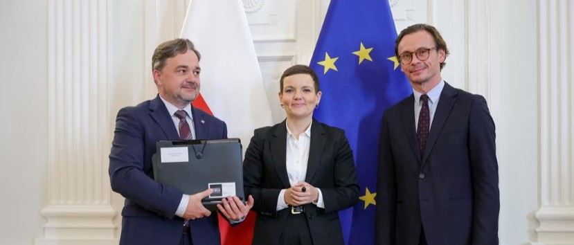 Trzy osoby w formalnych garniturach stoją na tle flagi Polski i flagi Unii Europejskiej w jasnym, ozdobnym wnętrzu; mężczyzna po lewej trzyma laptop z nalepkami, kobieta na środku ma złożone dłonie, a mężczyzna po prawej ma okulary.