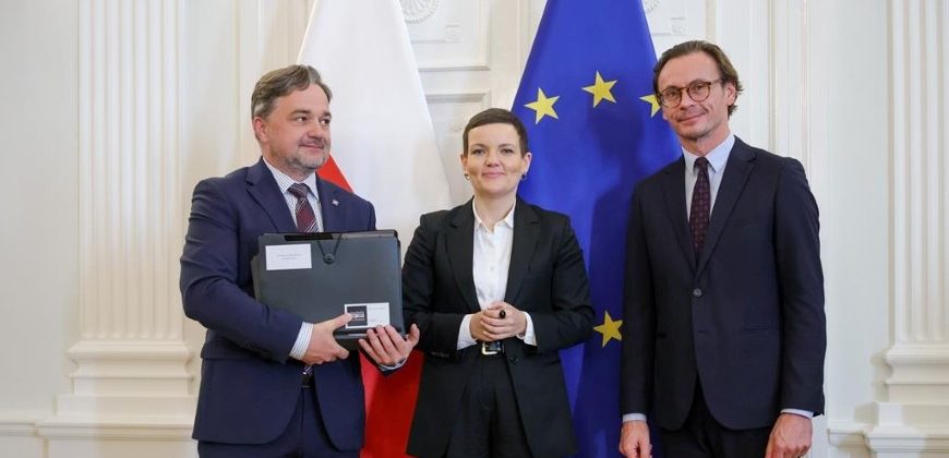 Trzy osoby w formalnych garniturach stoją obok siebie w jasnym, ozdobnym wnętrzu przed flagami Polski (biało-czerwoną) i Unii Europejskiej (niebieską z żółtymi gwiazdami); osoba po lewej trzyma zamknięty laptop, pośrodku stoi kobieta, po prawej mężczyzna w okularach.