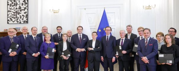 Grupa około dwudziestu formalnie ubranych osób ustawionych w rzędzie w eleganckim białym wnętrzu z drewnianym parkietem, pośrodku za nimi widoczne flaga Polski i flaga Unii Europejskiej oraz umieszczony nad drzwiami herb Polski; część osób trzyma czarne teczki z białymi etykietami, po lewej stronie na ścianie abstrakcyjny czarno-biały obraz i regał z książkami.