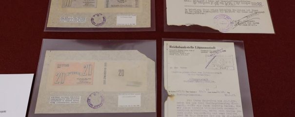 Na ciemnoczerwonym tle, ułożone w przezroczystych koszulkach, cztery pożółkłe archiwalne materiały: u góry po lewej arkusz z dwoma banknotami opatrzonymi cyfrą 