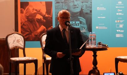 Mężczyzna w garniturze i okularach stoi przy mikrofonie czytając z teczki na scenie przed kolorowym banerem z dużymi portretami i logotypami, obok drewniane krzesła i okrągły stolik z butelkami wody oraz widoczny ekran kamery w prawym dolnym rogu.