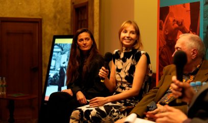 Trzech panelistów siedzi w rzędzie na scenie: pośrodku, nieco po prawej, młoda blondynka uśmiechnięta trzyma mikrofon i ma czarno-białą wzorzystą sukienkę, z lewej kobieta z długimi ciemnymi włosami w ciemnym ubraniu patrzy w stronę rozmówczyni, po prawej na pierwszym planie częściowo widoczny siwy mężczyzna z mikrofonem; w tle ekran z czarno-białym zdjęciem, kolorowy plakat z czerwoną fotografią, drewniane drzwi i ciepłe, pomarańczowe oświetlenie.