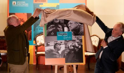 Dwaj mężczyźni odsłaniają połyskującą beżową tkaniną plakat ustawiony na sztaludze, na którym widać czarno-białe zdjęcia i napis Wajda; mężczyzna po prawej trzyma mikrofon, w tle kolorowy baner z napisem ARCHIWA PAŃSTWOWE i WYSTAWA oraz widoczne krzesła.