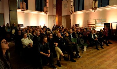 Wnętrze dużej, ciepło oświetlonej sali z wieloma osobami siedzącymi w rzędach krzeseł, wysokimi drewnianymi filarami i ozdobnymi żyrandolami, po prawej regał z napojami i napisem 